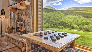 A stone fireplace with a log shelf, a chessboard, and a view of a forested mountain range.