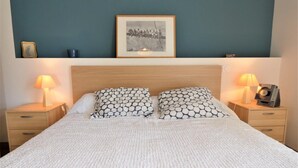 Ein ordentlich bezogenes Bett mit weißer, strukturierter Bettdecke, zwei gemusterten Kissen und einem gerahmten Bild darüber. Auf einem Nachttisch stehen eine Lampe und ein Radio.