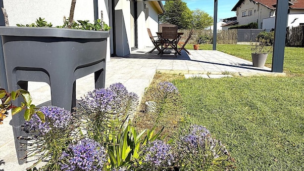 Eine überdachte Terrasse mit Tisch und Stühlen, ein grünes Rasenstück und ein Gebäude im Hintergrund.