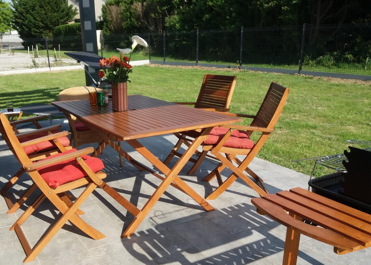 Eine Terrasse im Freien mit einem Holztisch und vier Klappstühlen, einem kleinen Tisch mit Getränken und einer Blumenvase sowie einem Grill.
