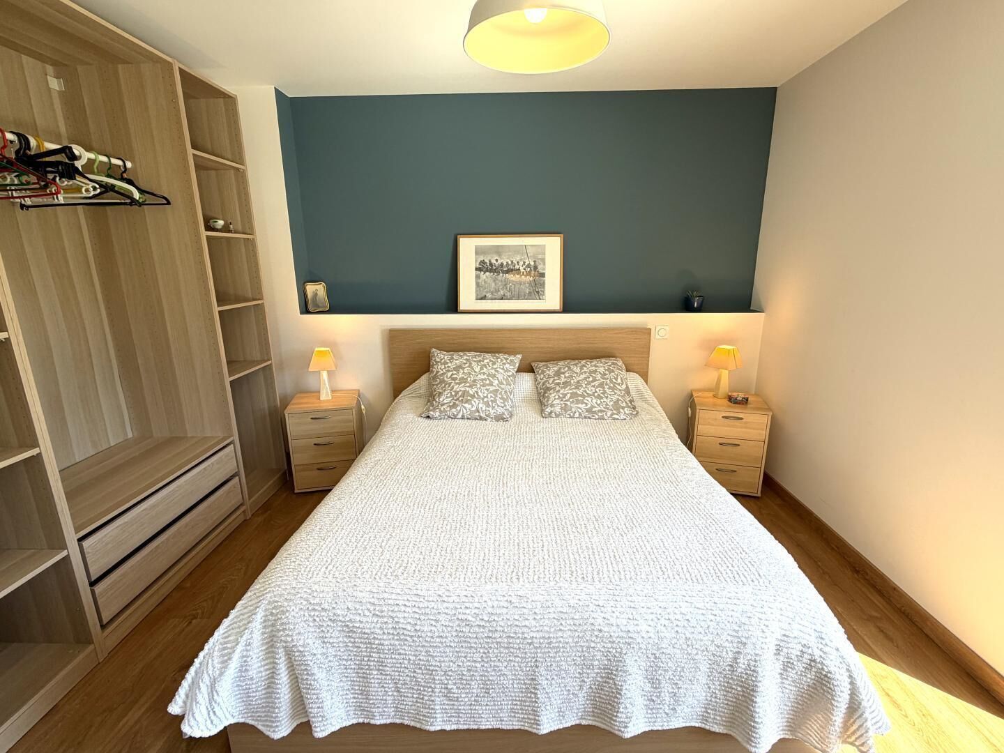 A bedroom with a bed, two bedside tables, a wardrobe, a framed picture on the wall, and a ceiling light.