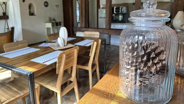 Un coin repas rustique avec des tables et des chaises en bois, un bocal rempli de pommes de pin posé sur une table, et une cuisine en arrière-plan.