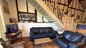 Un salon avec un canapé en cuir bleu foncé, une chaise en bois, un téléviseur et un escalier dont les poutres en bois sont apparentes.