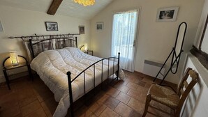 Une chambre à coucher comprenant un lit, deux tables de chevet avec des lampes, une chaise en bois, une fenêtre avec des rideaux et un luminaire au plafond.