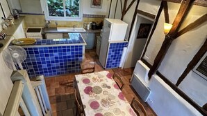 Une cuisine aux carreaux bleus, une table à manger recouverte d’une nappe à motifs et une fenêtre donnant sur un paysage verdoyant.