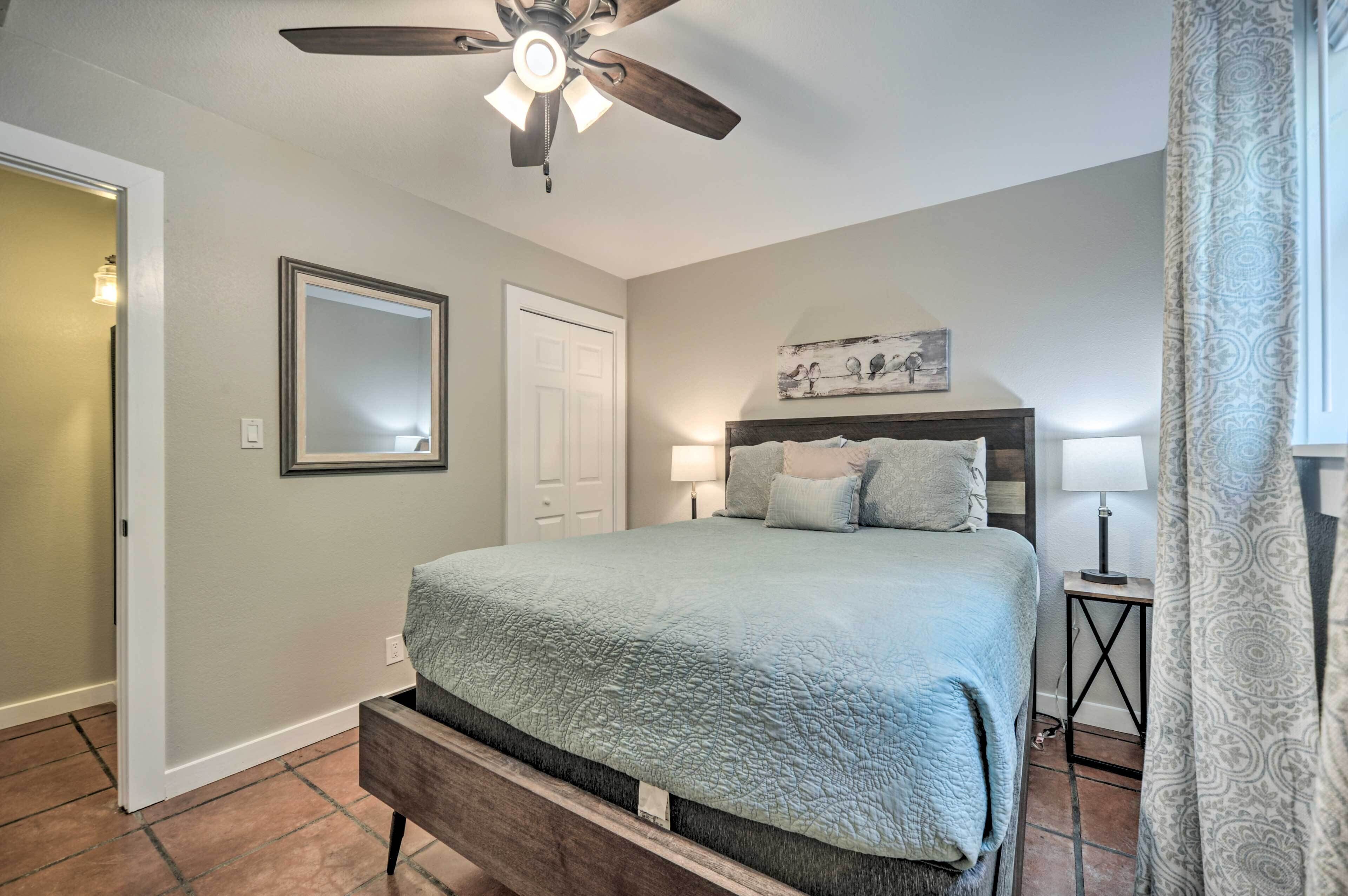 A bedroom with a bed, a ceiling fan, a nightstand, a lamp, a mirror, and a door.
