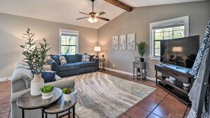 A living room with a sofa, coffee table, and TV stand. There are plants, a ceiling fan, and a window.