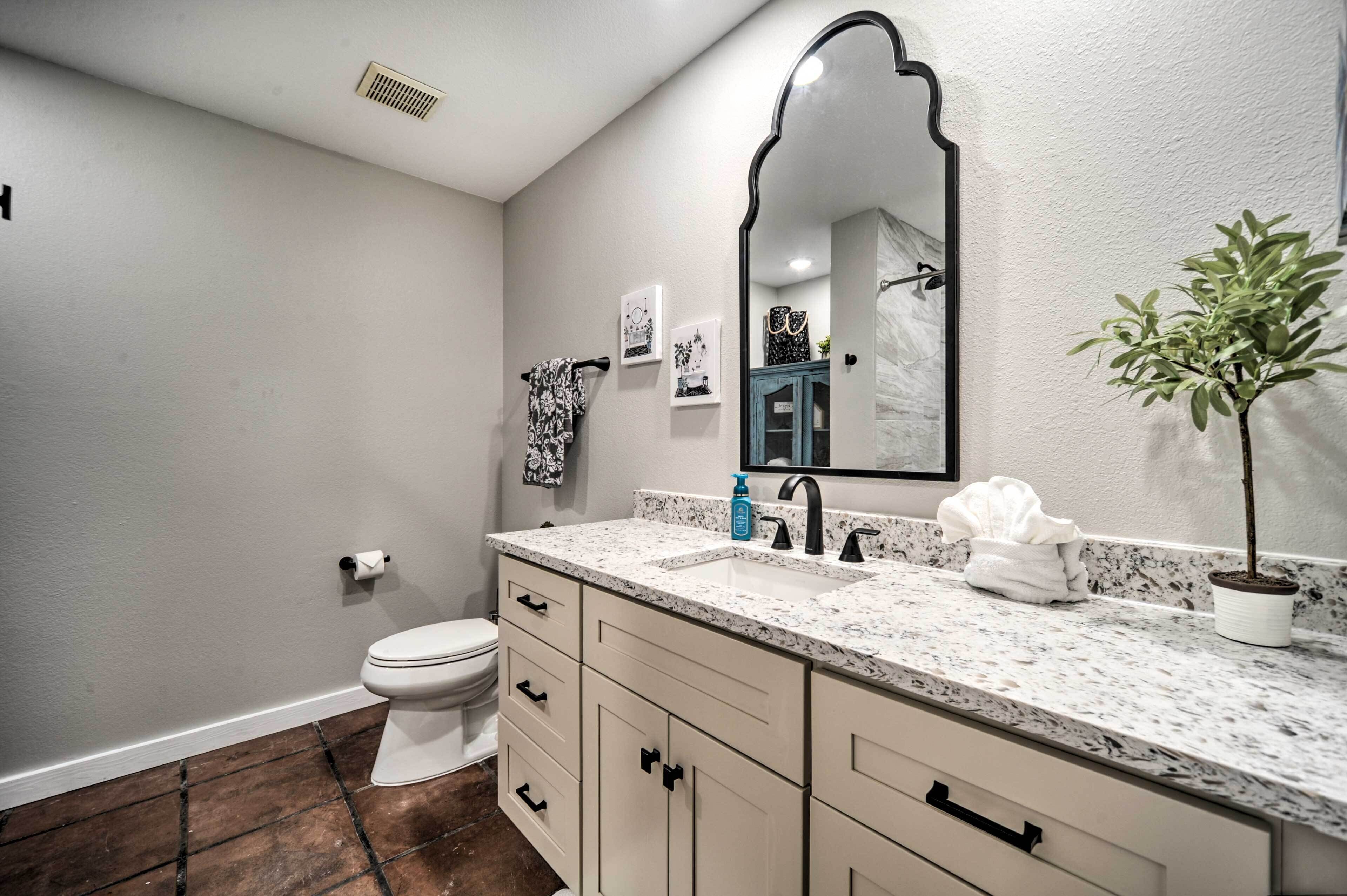 Un baño con inodoro blanco, lavabo con encimera de mármol, espejo y planta en maceta.