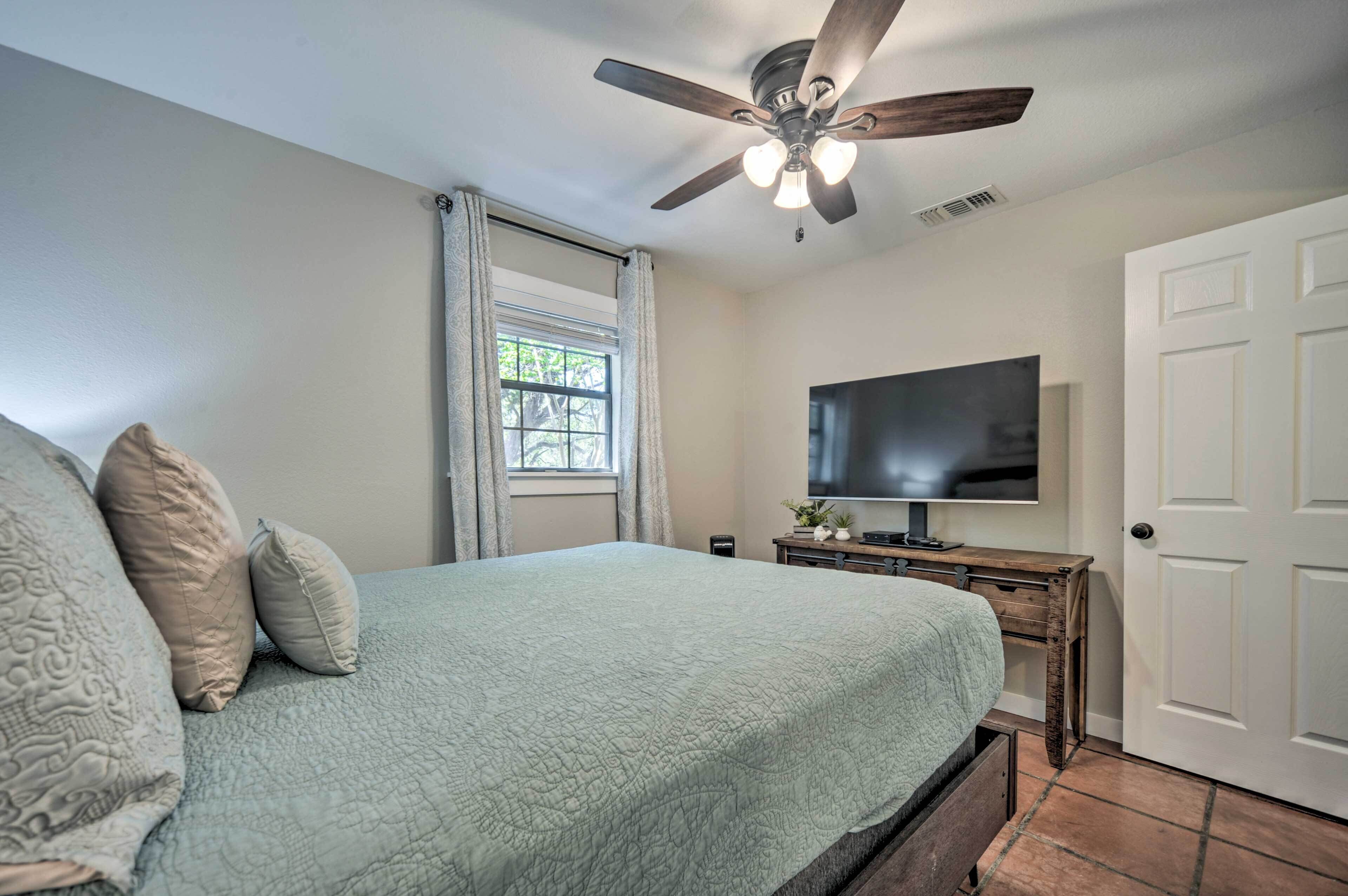 A bedroom with a bed, a ceiling fan, a TV, a window with curtains, and a door.