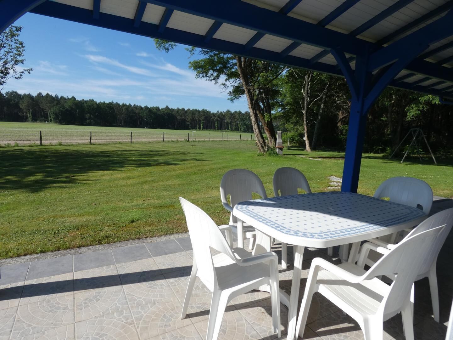 Une terrasse extérieure avec une table et des chaises, un espace abrité et une vue sur un champ verdoyant et des arbres.