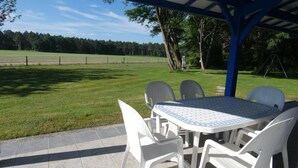 An outdoor patio with a table and chairs, a covered area, and a view of a grassy field and trees. - Gîte de Deze - Herm - Gîte (HERM)