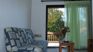 A room with a patterned sofa, a small wooden table, and a view of greenery through a sliding glass door. - Tamaris - Morcenx - Gîte (MORCENX)