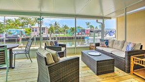 Un espacio de estar al aire libre con techo, amueblado con mimbre, una mesa y vistas a un puerto deportivo con barcos y edificios.