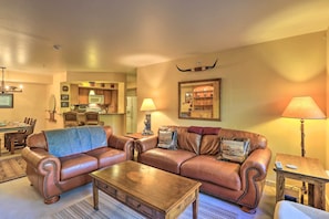 A living room with a brown leather sofa, a wooden coffee table, and a lamp. There is a kitchen in the background with a microwave and a refrigerator. - Walkable Dtwn Condo ~ 5 Miles to Beaver Creek Ski! (Edwards)