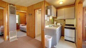 A compact kitchen with a stove, sink, and cabinets. There is a small bathroom with a shower and toilet. A bed is visible in the background. - Convenient Cabin w/ Access to 3 Boat Docks! (Stone Lake)