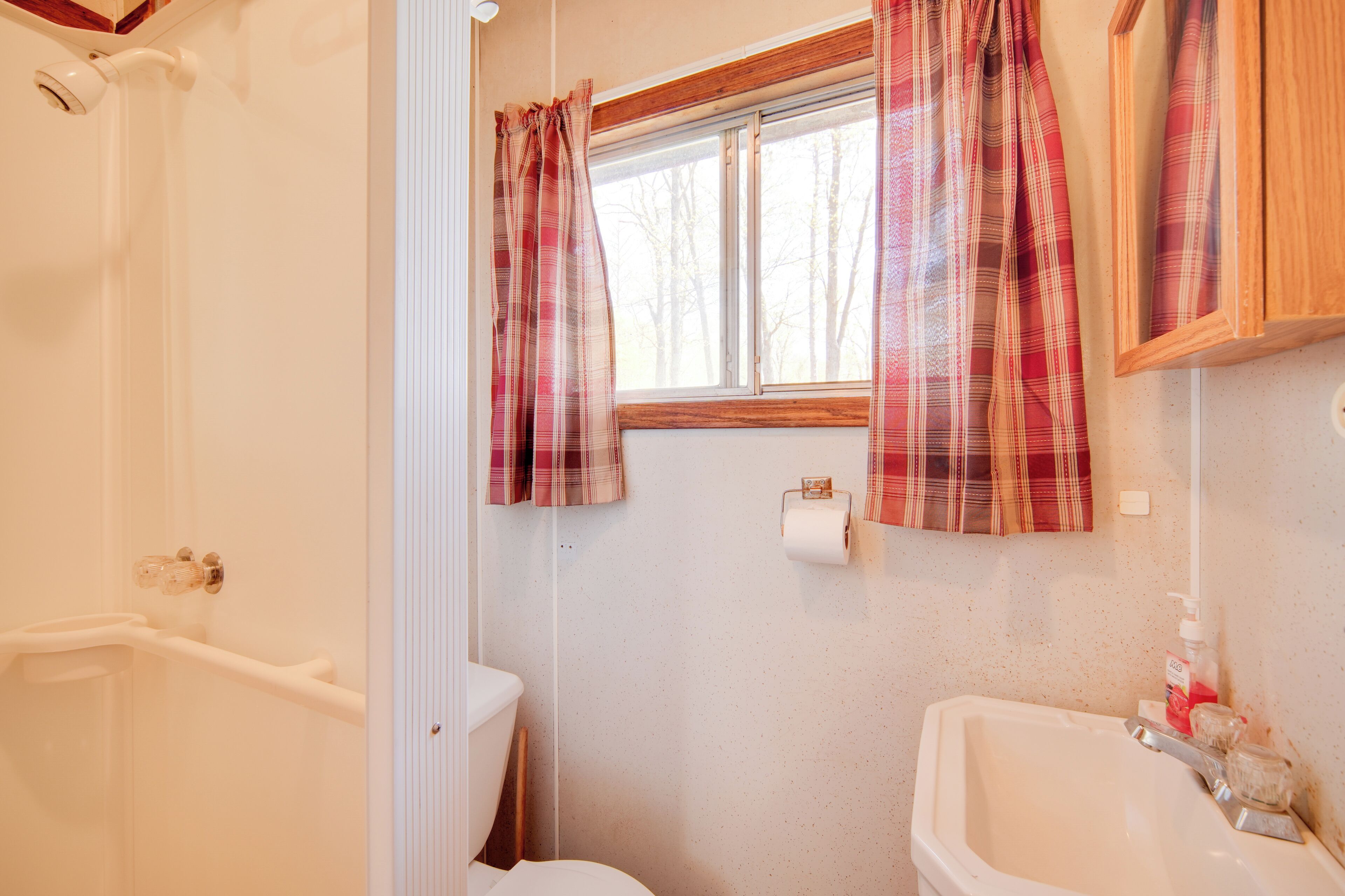 A bathroom with a toilet, sink, and shower. There is a window with curtains and a mirror above the sink.