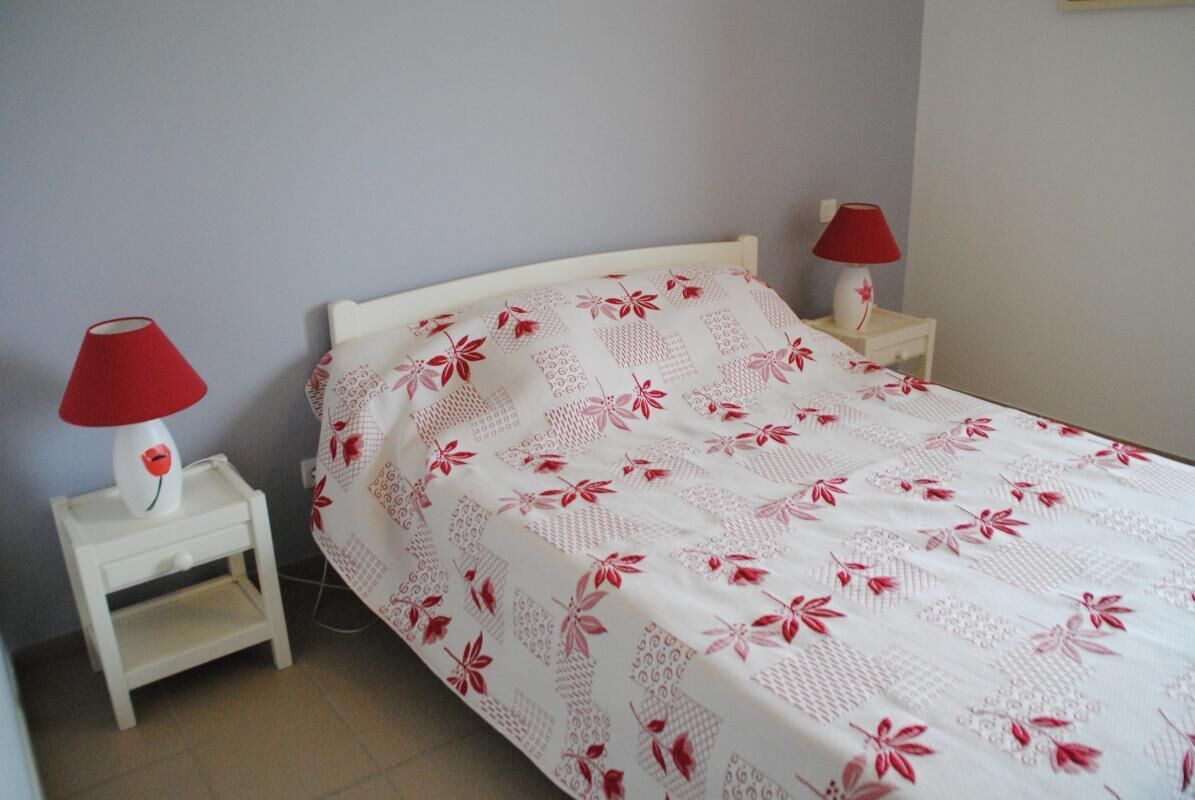 A bedroom with a white bedspread featuring red floral patterns, two bedside tables with red lamps, and a white nightstand.