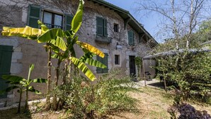 Un bâtiment rustique en pierre, doté de volets verts, un jardin planté de bananiers et un ciel dégagé.