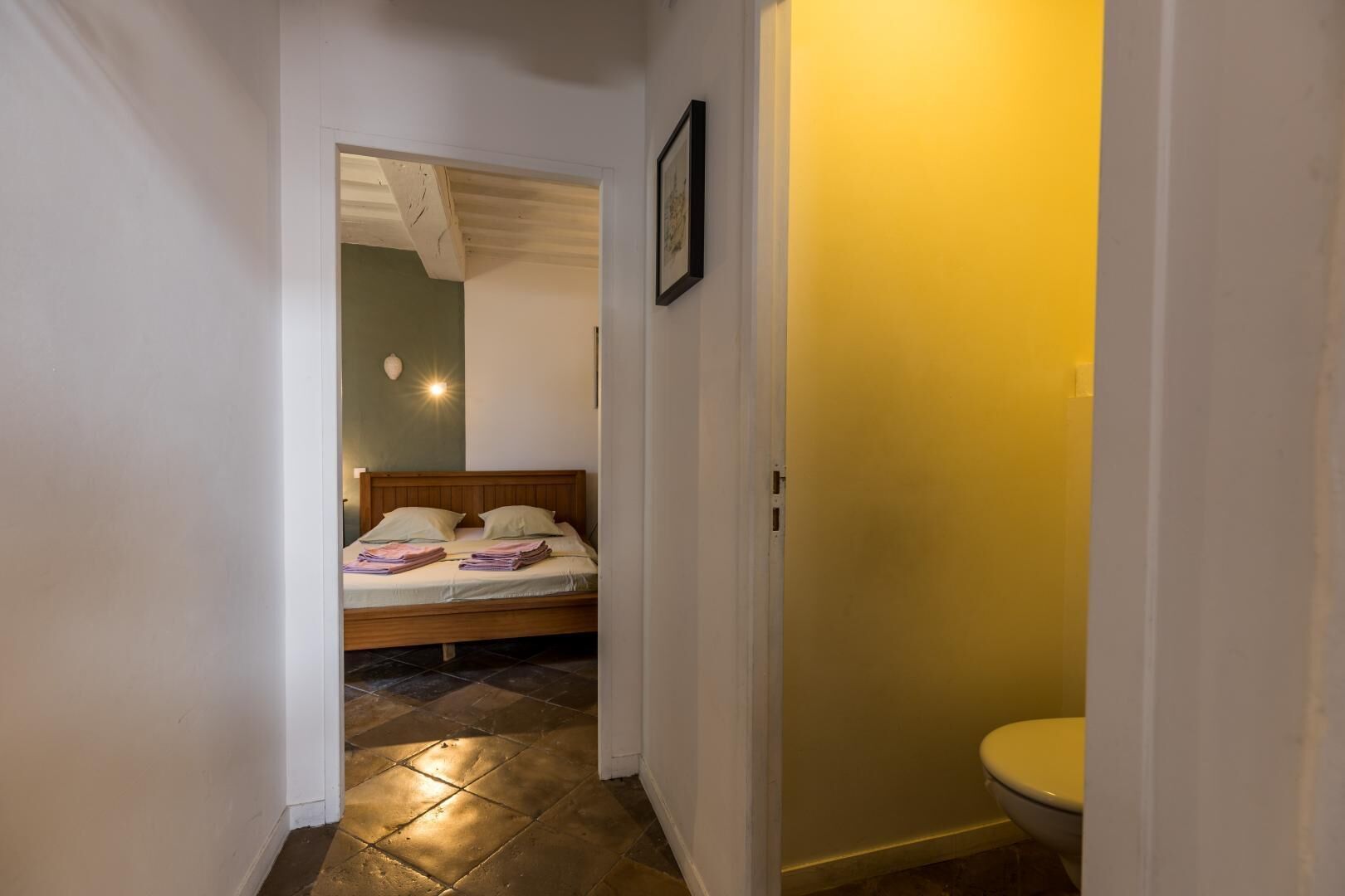 A narrow hallway leading to a bedroom with a bed, a bedside lamp, and a framed picture on the wall.