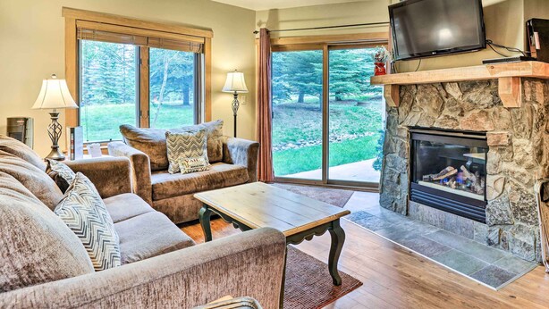 A living room with a stone fireplace, a television mounted above it, and a large window offering a view of the outdoors. - Cozy Red Hawk Lodge Condo: Walk to River Run! (Keystone)