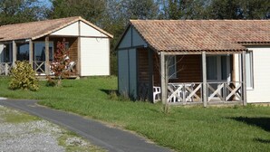 Two wooden cabins with tiled roofs, a gravel path, and a grassy area with trees in the background.