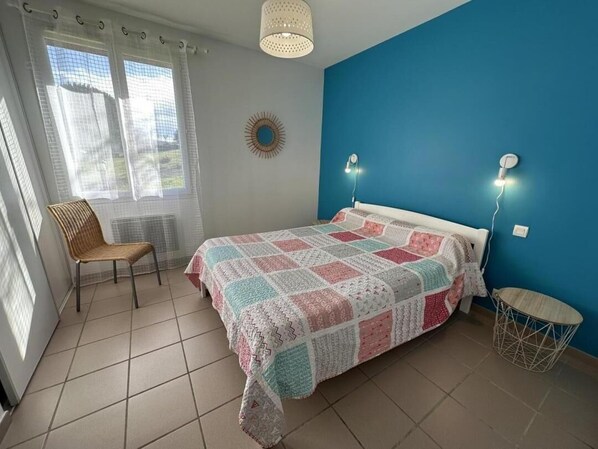 A bedroom with a bed, a chair, a small table, and a wall-mounted lamp.
