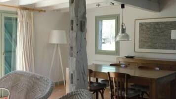 Une pièce spacieuse avec de hauts plafonds et des poutres apparentes, comprenant un coin repas avec une table et des chaises en bois, des fauteuils en osier et une grande fenêtre laissant entrer la lumière naturelle.