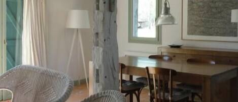 Amplia habitación con techos altos y vigas a la vista, que incluye una zona de comedor con mesa y sillas de madera, sillas de mimbre y un ventanal que proporciona luz natural.