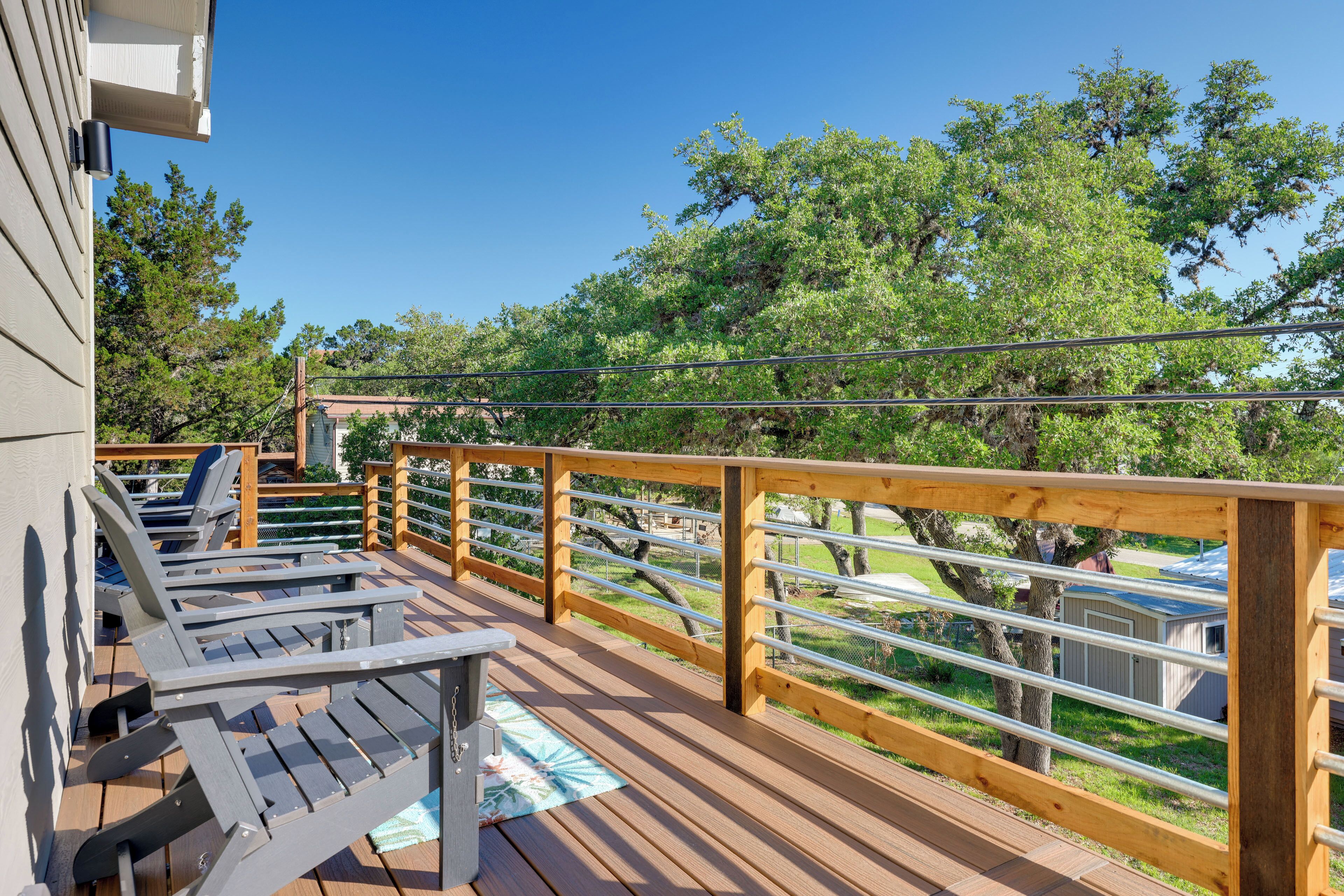 Terraza de madera con sillones de descanso y una mesa, con vista a un área verde con árboles y un edificio.