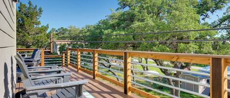 Terraza de madera con sillones de descanso y una mesa, con vista a un área verde con árboles y un edificio.