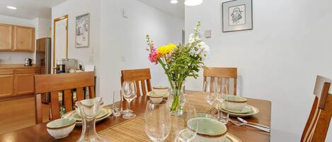 A dining area with a wooden table set for four, surrounded by chairs. There is a vase with flowers on the table, and the room has wooden cabinets and a white wall with framed pictures.