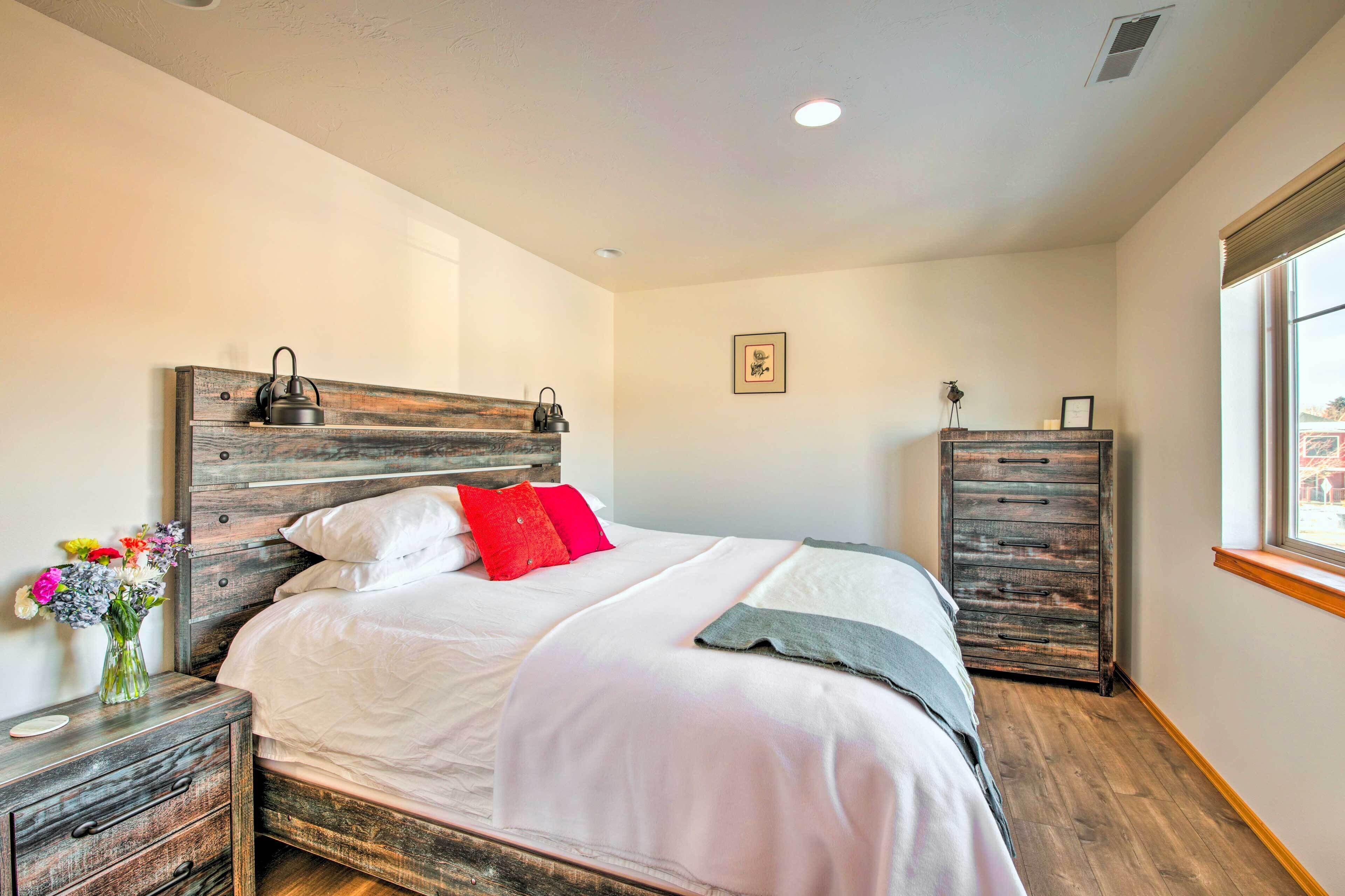 Une chambre à coucher bien aménagée, avec une tête de lit en bois, une table de chevet ornée d’un vase de fleurs, une commode et une fenêtre offrant une vue agréable.