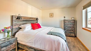 Une chambre à coucher bien aménagée, avec une tête de lit en bois, une table de chevet ornée d’un vase de fleurs, une commode et une fenêtre offrant une vue agréable.