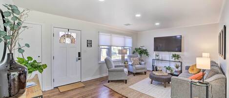 Una sala de estar moderna con un sofá, sillones, una mesa de centro y un televisor. Se observa una puerta, una ventana con persianas y una planta sobre una mesa.