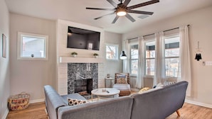 A modern living room with a fireplace, a ceiling fan, and a large window with curtains. - Charming Downtown Home w/ Updated Interior! (Oklahoma City)