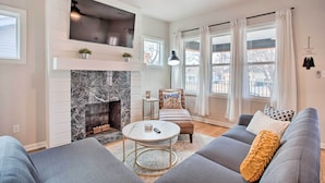 A modern living room with a fireplace, a ceiling fan, and a large window with curtains. - Charming Downtown Home w/ Updated Interior! (Oklahoma City)