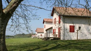 Une rangée de bâtiments blancs aux volets rouges et aux toits en tuiles, situés dans un champ verdoyant avec un arbre au premier plan.