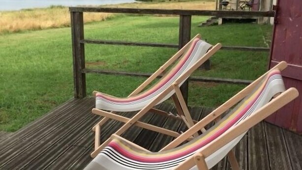 Deux fauteuils de jardin à rayures, placés sur une terrasse en bois, offrent une vue sur un lac et des prairies verdoyantes.