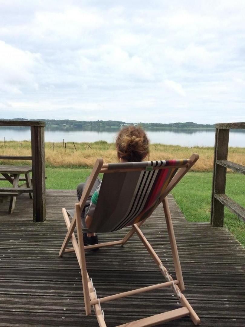Iemand zit op een houten terrasstoel, uitkijkend over een meer, met een picknicktafel en een gazon op de voorgrond.