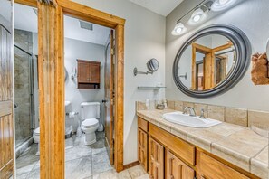 A bathroom with a wooden vanity, a round mirror, and a sink. There is a shower area with a glass door and a toilet visible in the background. - Ruidoso Condo w/ Hot Tub: Long Walk to Midtown! (Ruidoso)