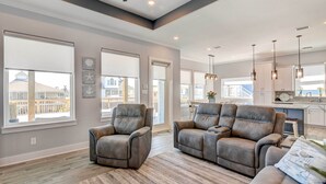 A spacious living room with a gray sofa and armchairs, large windows, and a modern kitchen in the background.