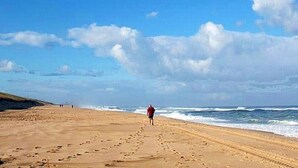 Ein Strand mit Fußspuren, die zum Wasser führen, eine Person geht spazieren, und ein klarer Himmel.