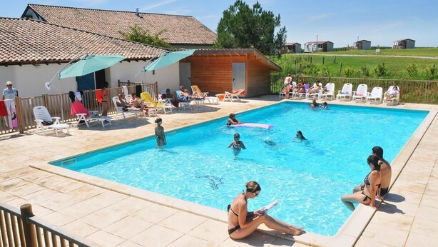 Piscina com pessoas nadando e relaxando. Há espreguiçadeiras e um edifício de madeira ao fundo.