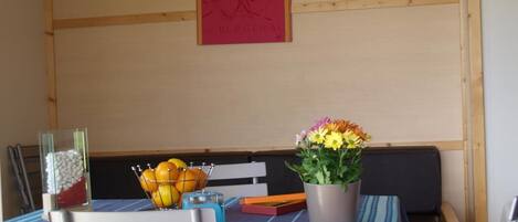 A dining area with a table set for two, featuring a striped tablecloth, a vase with flowers, and a bowl of oranges.