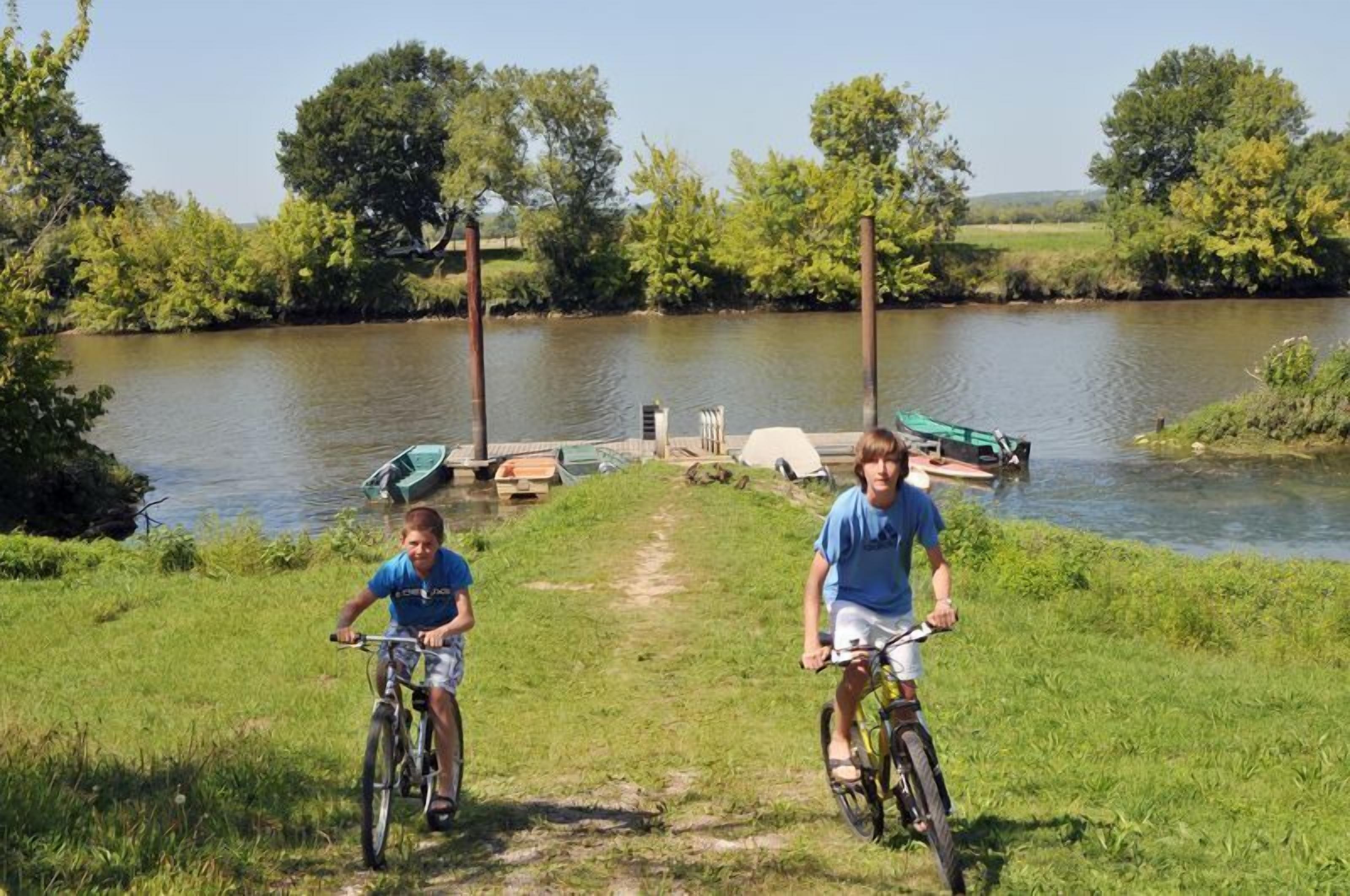 Two people riding bicycles on a grassy path beside a calm river with boats docked at a pier.