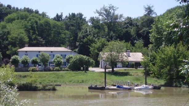 Un lago tranquillo con barche ormeggiate, circondato da una vegetazione rigogliosa e due case con finestre e tetti ben visibili.