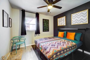 Une chambre à coucher avec un lit, un ventilateur de plafond, une fenêtre avec des rideaux et des œuvres d’art encadrées accrochées aux murs.