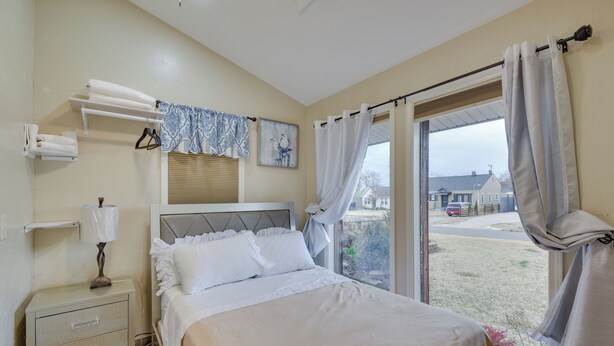 A bedroom with a bed, a nightstand, a lamp, a window with curtains, and a view of the outside. - Cozy Oklahoma City Home w/ Deck & Gas Grill! (Oklahoma City)