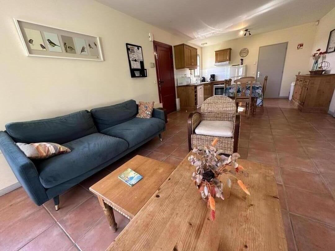 Un salon avec un canapé, une table basse et un fauteuil. On aperçoit une cuisine au fond.