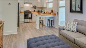 Una cocina moderna con armarios blancos, una isla central y un comedor con taburetes. Hay un otoman tapizado en gris y un sofá con un cojín estampado.